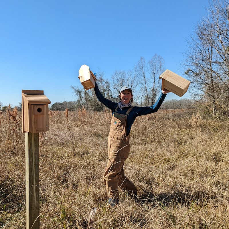 In the image, a person is joyfully holding up two wooden birdhouses in a field. They are standing next to a birdhouse mounted on a wooden post. The person is wearing overalls and a long-sleeved shirt, with a happy expression on their face. The background shows a clear blue sky and dry grass.